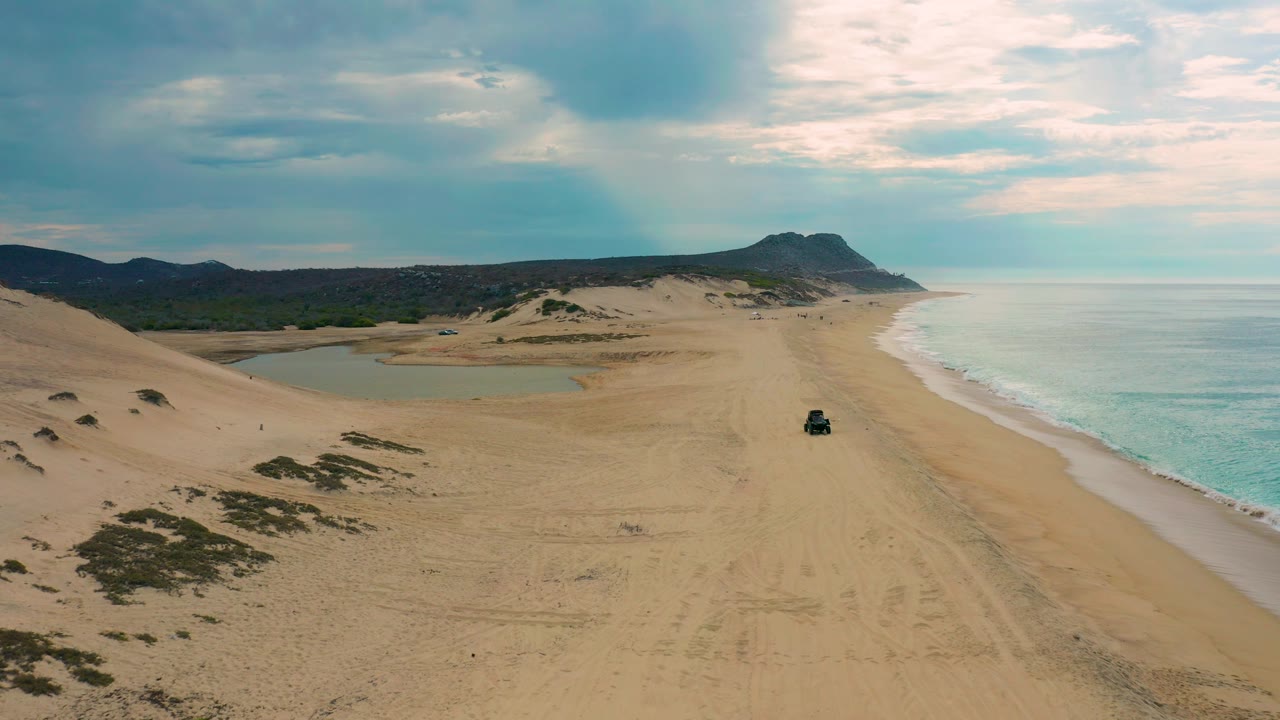 Laguna Beach aerial frame showing tire-marked sand in the foreground, low dunes and inland vegetation to the left, blue-green ocean to the right, distant headland, and no visible people.
