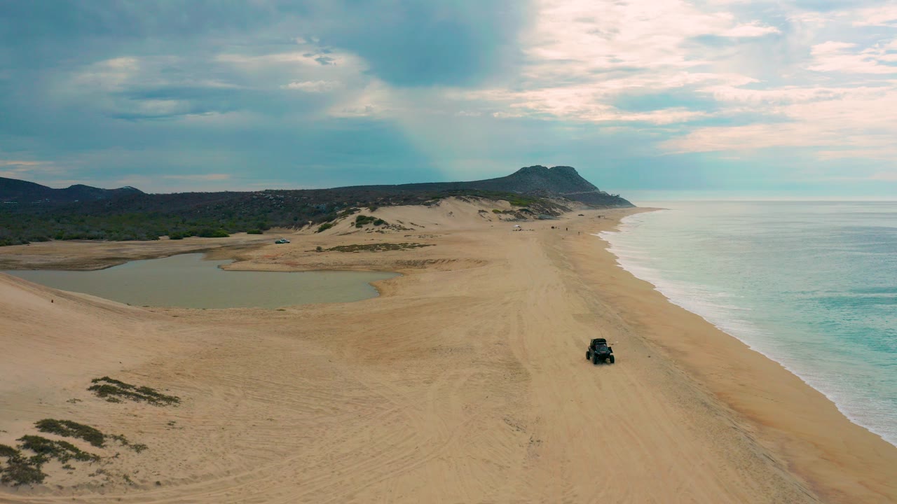 Aerial view over Laguna Beach in Baja California Sur, Mexico, showing a broad sandy beach, pale surf on the right, lagoon water on the left, distant hills, a small dark vehicle, and no visible people.