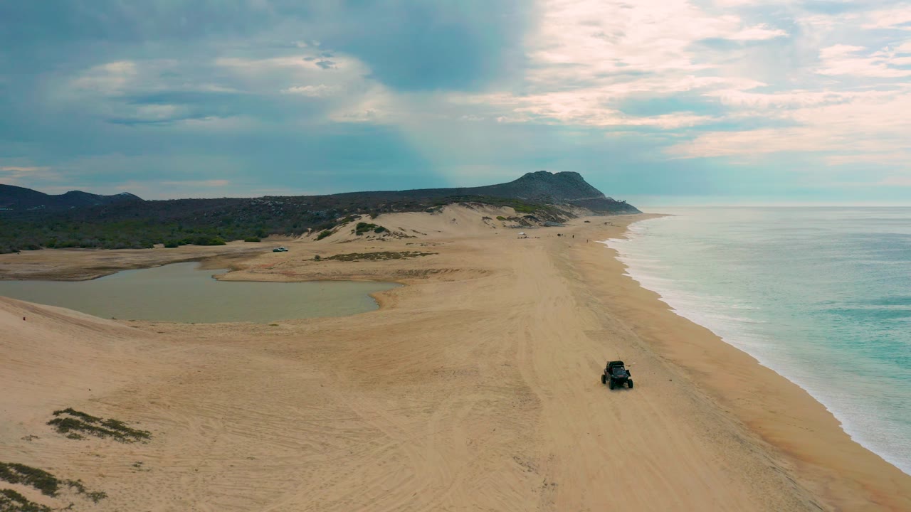 Wide aerial frame from Laguna Beach, Baja California Sur, Mexico, showing the vehicle closer to the lower right sand, surf washing the right shoreline, lagoon water left of center, low dunes, distant hills, and no visible people.