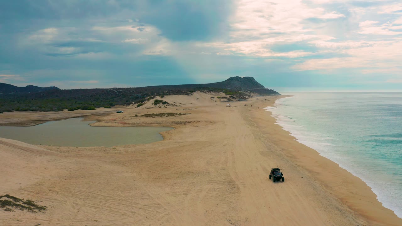 High drone frame over Laguna Beach in Baja California Sur, Mexico, with lagoon water prominent on the left, a long sandy beach running toward distant hills, white surf on the right, a small vehicle near the lower right, and no visible people.