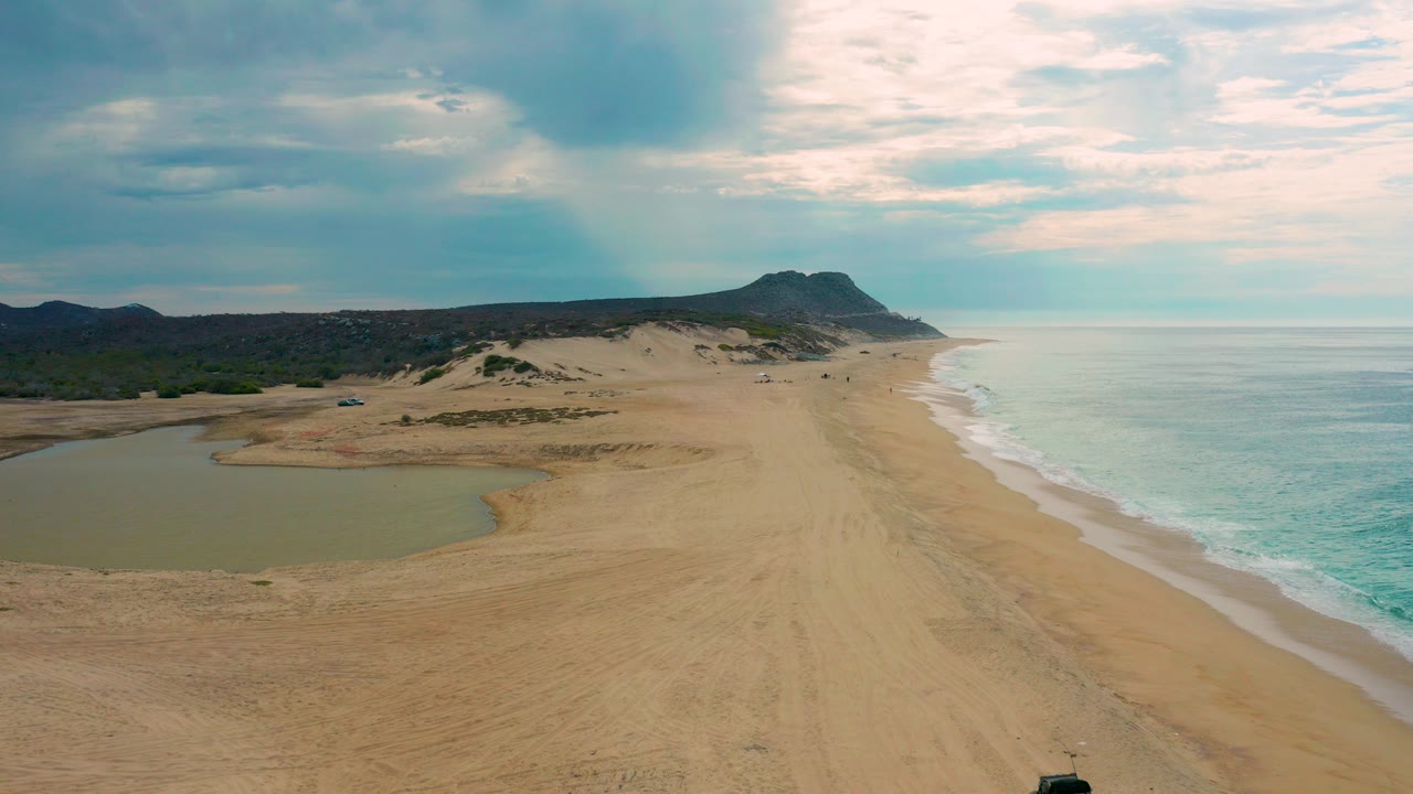 Aerial frame over Laguna Beach, Baja California Sur showing a sandy beach between a shallow lagoon and pale ocean surf, with dunes, a distant headland, cloud cover, and no visible people.