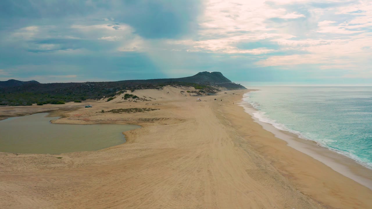 Elevated frame over Laguna Beach, Baja California Sur showing broad empty sand, turquoise ocean water, a shallow lagoon, scrubby dune areas, a low headland on the horizon, and no visible people.
