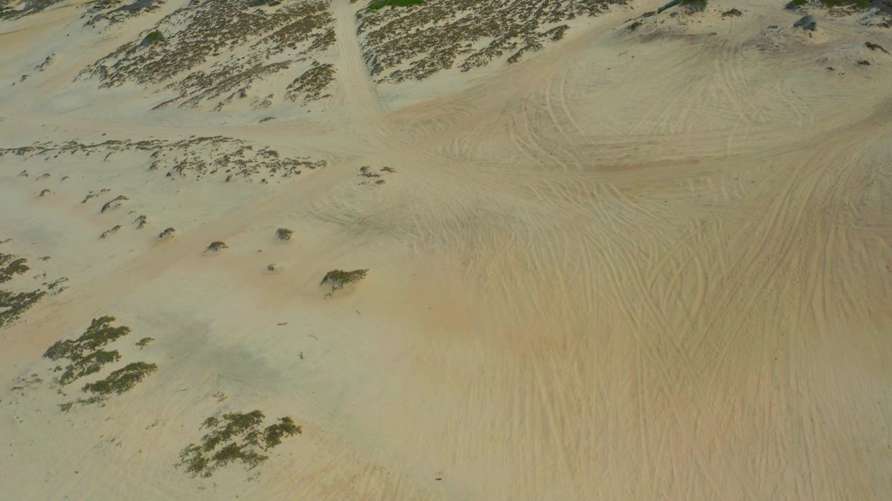 Aerial frame over Laguna Beach, Baja California Sur showing pale sand slopes, scattered dark scrub, faint track marks, and no visible people.