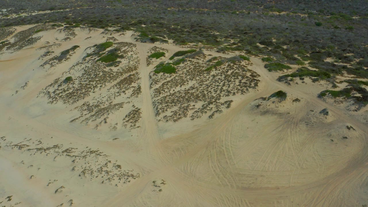 Aerial view over a shallow sandy basin in Laguna Beach, Baja California Sur, with scrub patches on the dunes, brushland at the top of frame, and no visible people.