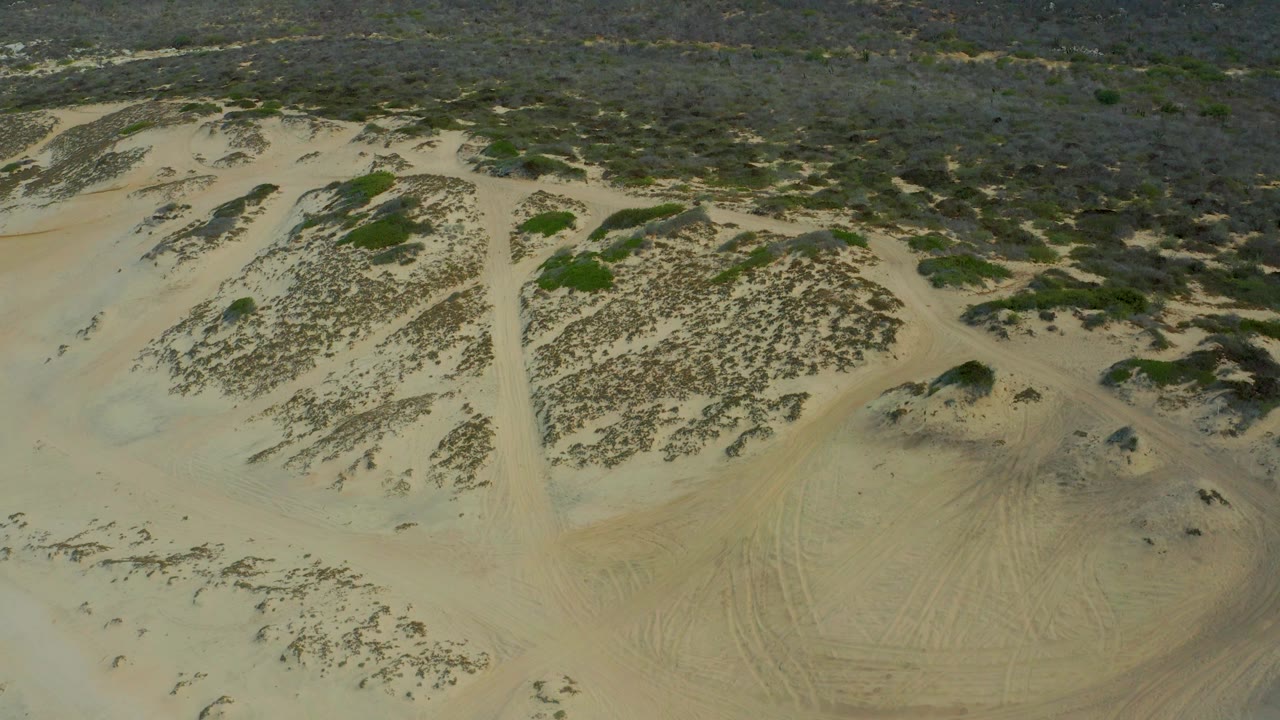 Mid-clip aerial frame in Laguna Beach, Baja California Sur with a narrow sandy track dividing scrub-covered dune mounds and no visible people.