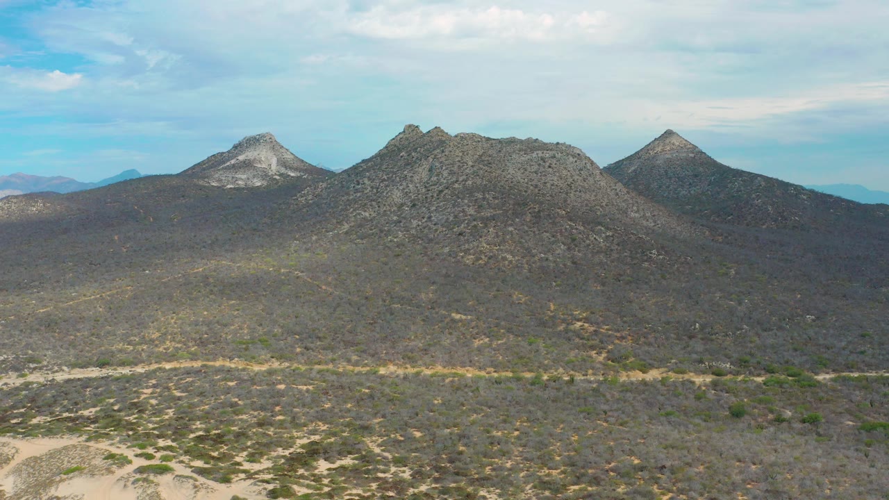 Aerial still over scrubby ground toward a rocky mountain ridge in Laguna Beach, Mexico, with scattered white clouds and no visible people.