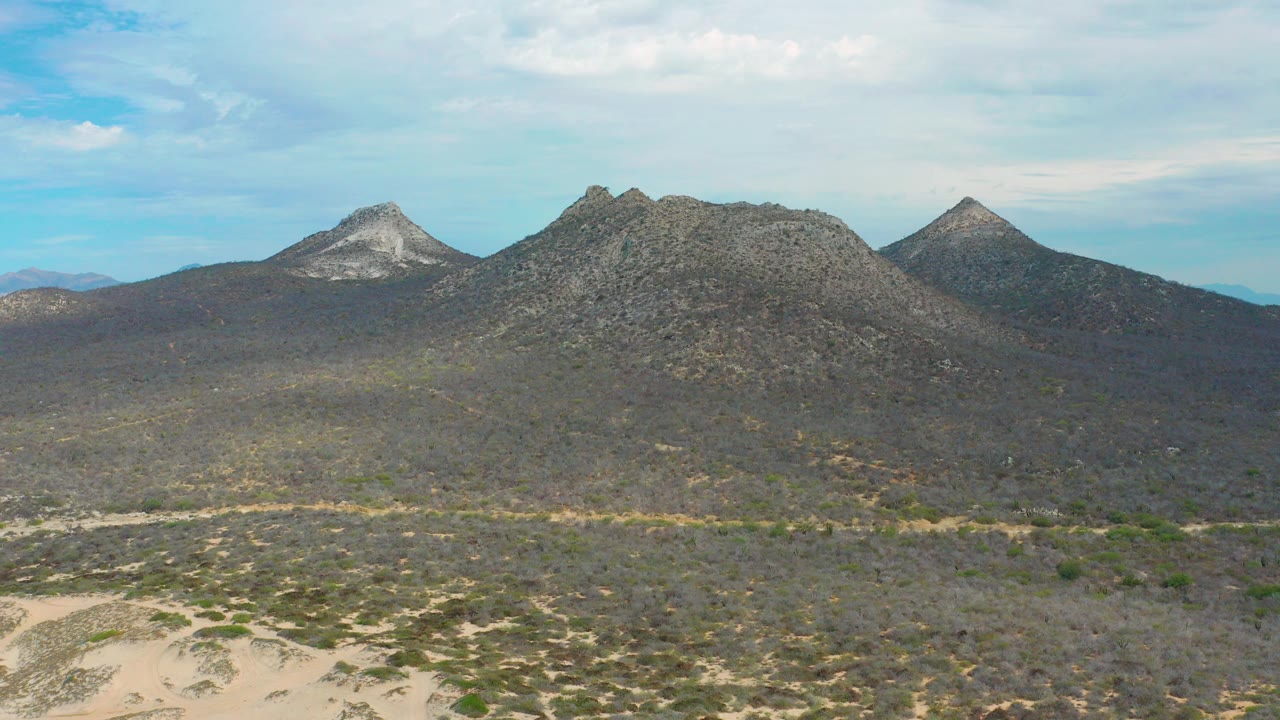 Opening frame of Laguna Beach, Baja California Sur, showing rocky mountain peaks beyond sandy scrubland under a blue sky with white clouds and no visible people.