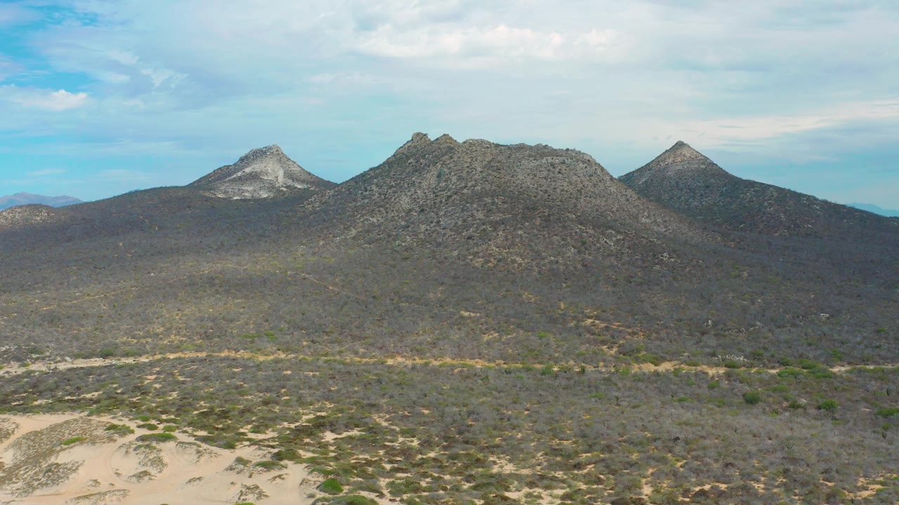 At 0:02, the frame shows three distinct mountain forms across the horizon, dark scrub and sandy patches below, and no visible people in the Laguna Beach, Baja California Sur landscape.