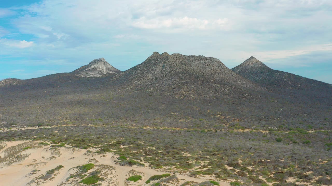 Opening frame of Laguna Beach, Baja California Sur terrain with pale dunes in front, low scrub across the flats, dark rocky mountains beyond, and no visible people.