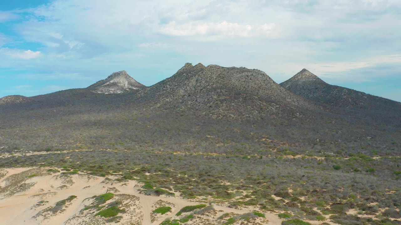 Laguna Beach, Baja California Sur view with the sandy foreground running across the lower frame, scrubland spreading toward three mountain forms, and no visible people.