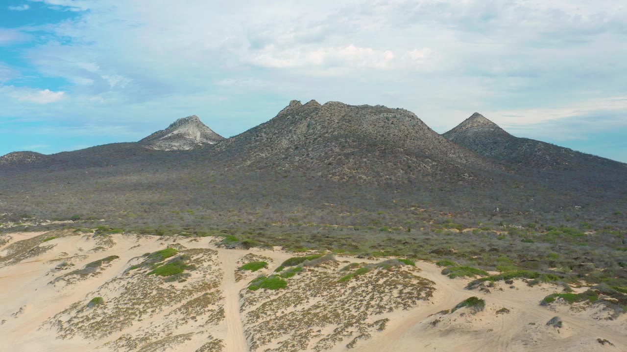 A broad aerial view in Laguna Beach, Baja California Sur, showing pale sand, scattered desert shrubs, a dark central hill mass, and lighter pointed peaks at left and right, no visible people.