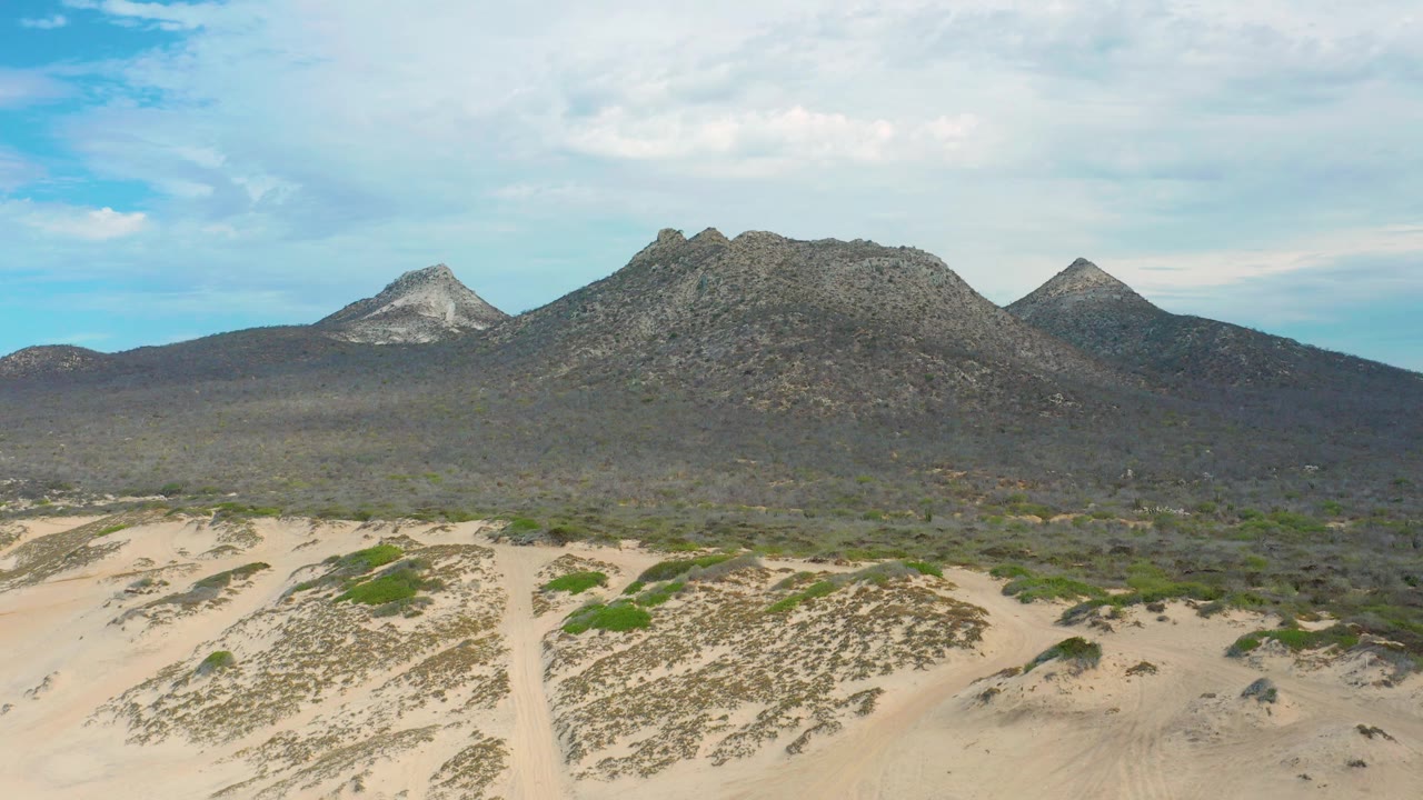 Mid-clip frame of Laguna Beach terrain with a lower band of sand, darker scrub across the plain, and rugged mountains set below a bright cloud layer, no visible people.