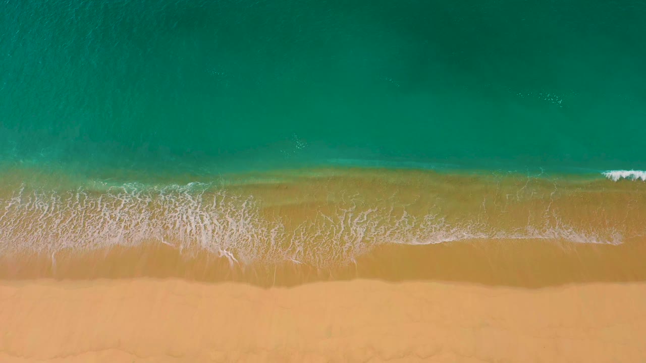 Overhead view of Laguna Beach in Baja California Sur where turquoise water meets a wide band of golden sand, with thin white foam and no visible people.
