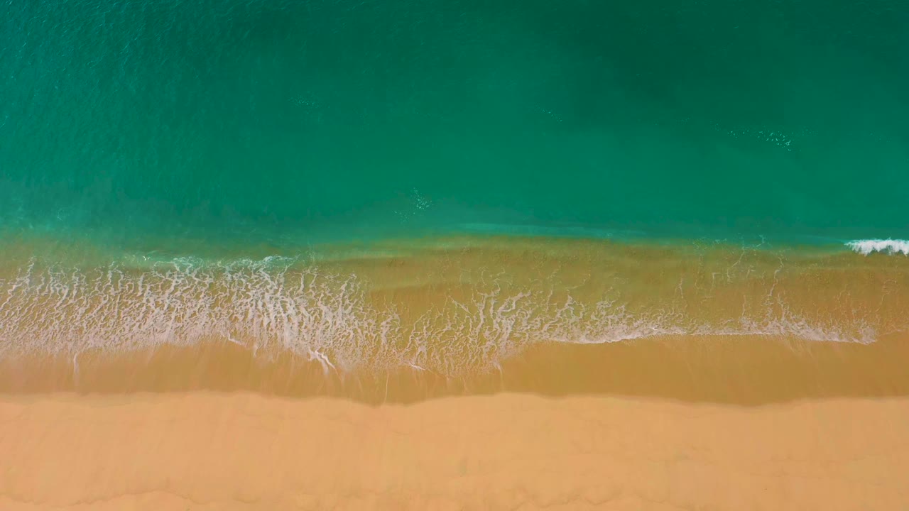 Clear blue-green water above a smooth sandy beach with light foam at the edge.