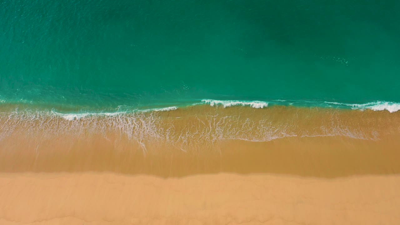 Overhead view of Laguna Beach in Baja California Sur with a longer white surf line spreading across the sand beneath clear teal water, with no visible people.