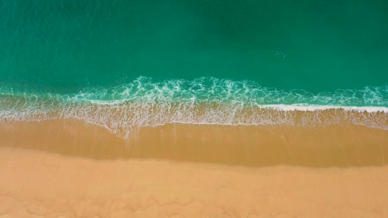A foaming wave spreads wider along the Laguna Beach shore, with green-blue water above the surf line, tan sand below, and no visible people.