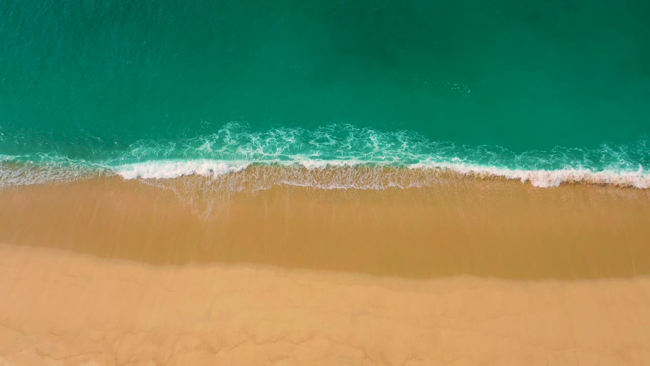 White surf curls along a mostly straight sandy shoreline in Baja California Sur, with darker green water offshore and no visible people.