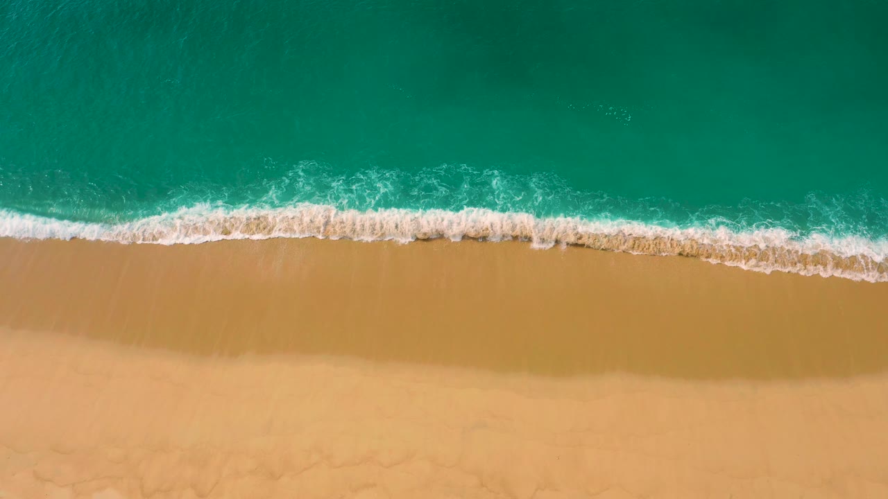 From above, a thicker band of white foam breaks onto empty tan sand at Laguna Beach, with clear green water filling the top of the frame and no visible people.