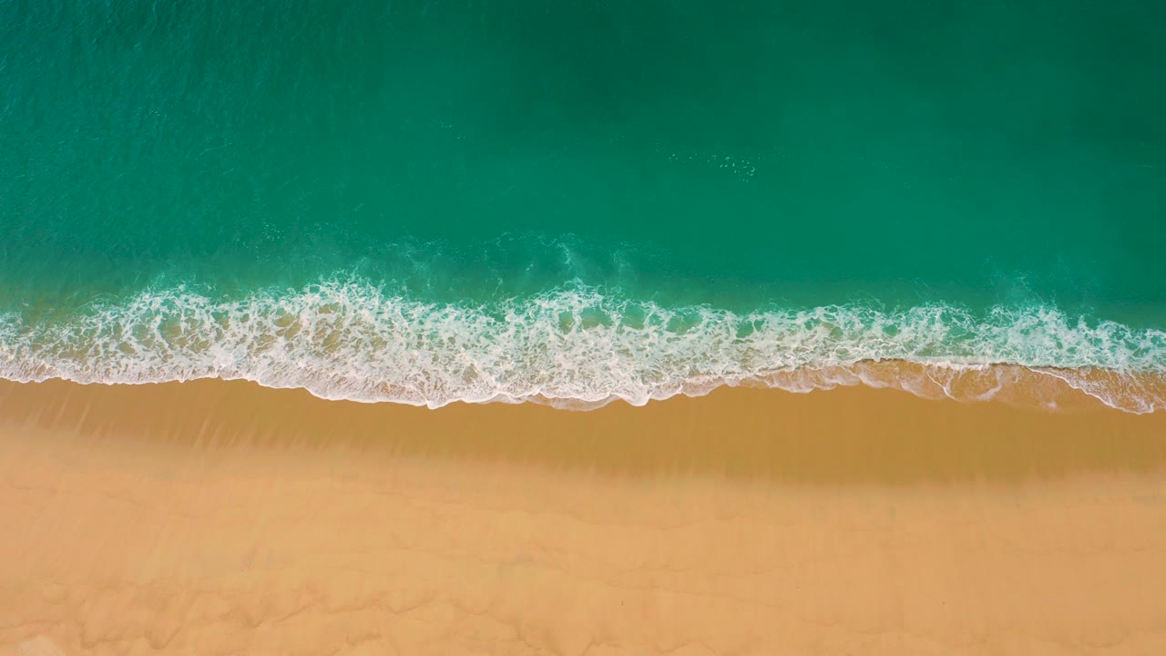 Overhead view of Laguna Beach, Baja California Sur, with turquoise water pushing a white foam line onto tan sand and no visible people.