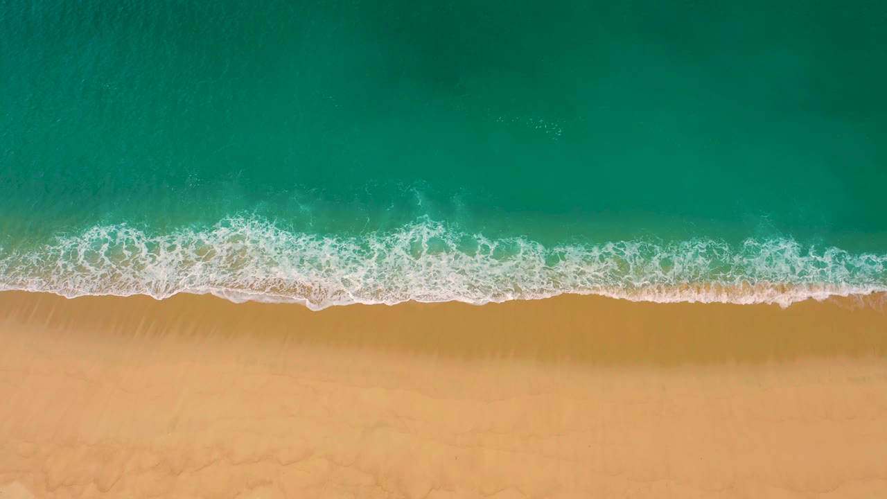 The overhead beach view shows a broken white wave edge running horizontally across Laguna Beach, Baja California Sur, with clear water above and smooth sand below, no visible people.