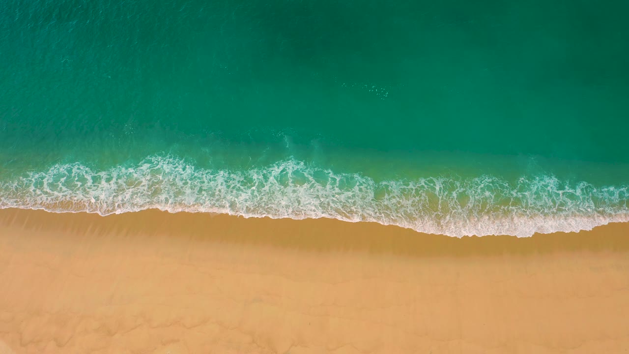 Turquoise surf washes into a thin white lace of foam over tan sand at Laguna Beach, Mexico, seen from directly above with no visible people or structures.