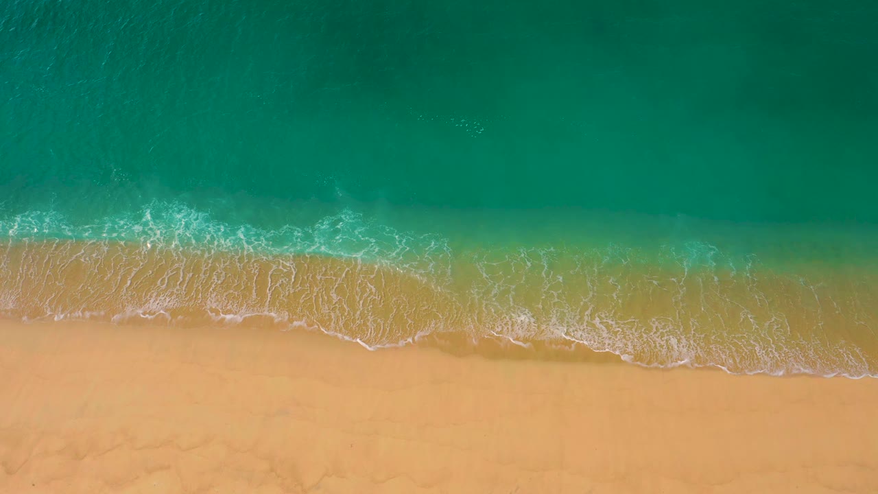 Aerial view at Laguna Beach, Baja California Sur, with a broader lace of white foam sliding over wet sand beneath clear green-blue water and no visible people.