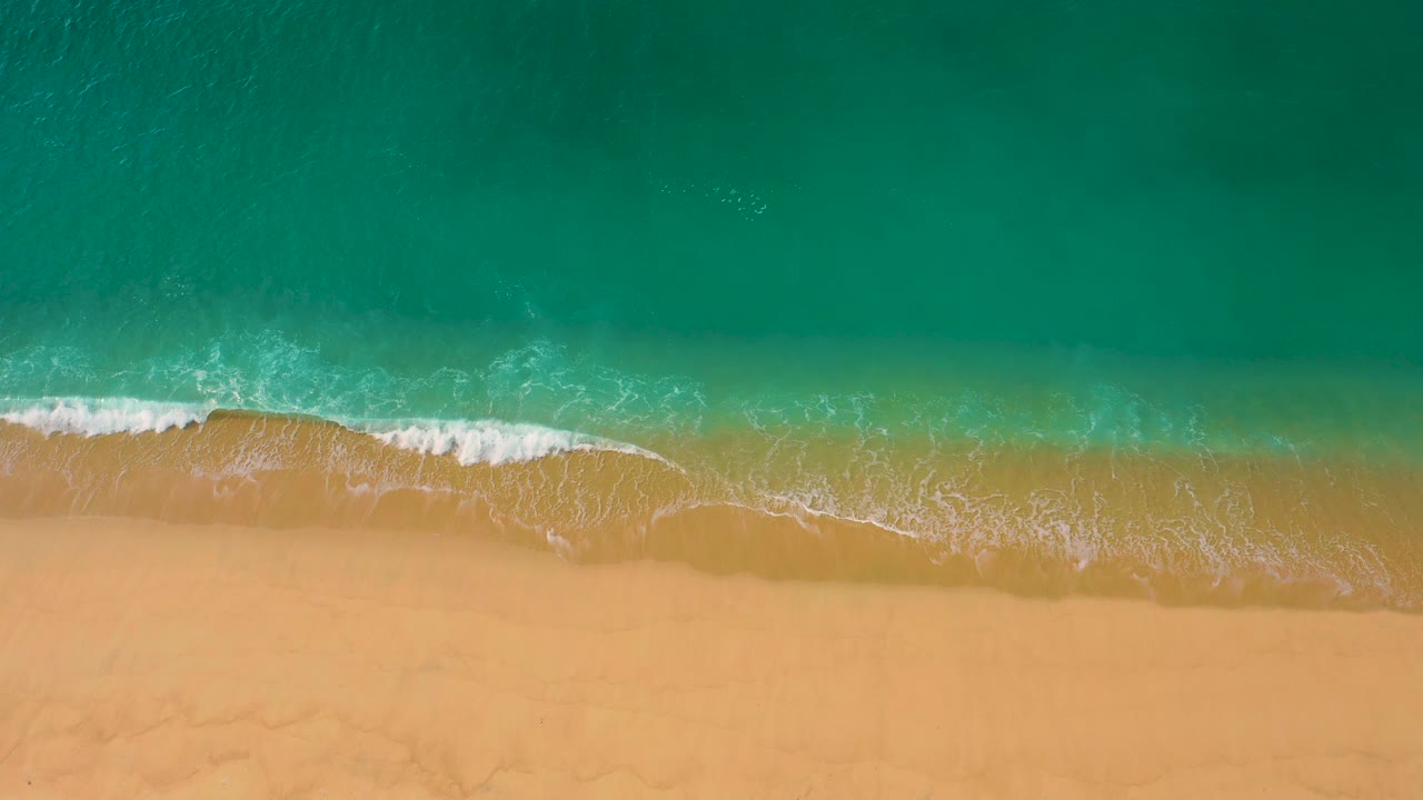 Top-down Laguna Beach shoreline frame with a small white breaker curving near the upper sand line, clear water above it, and no visible people.