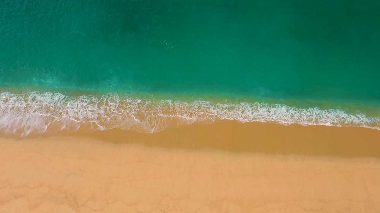 Aerial frame of white wave foam spreading across the yellow shoreline below emerald water at Laguna Beach, Baja California Sur, with no visible people.