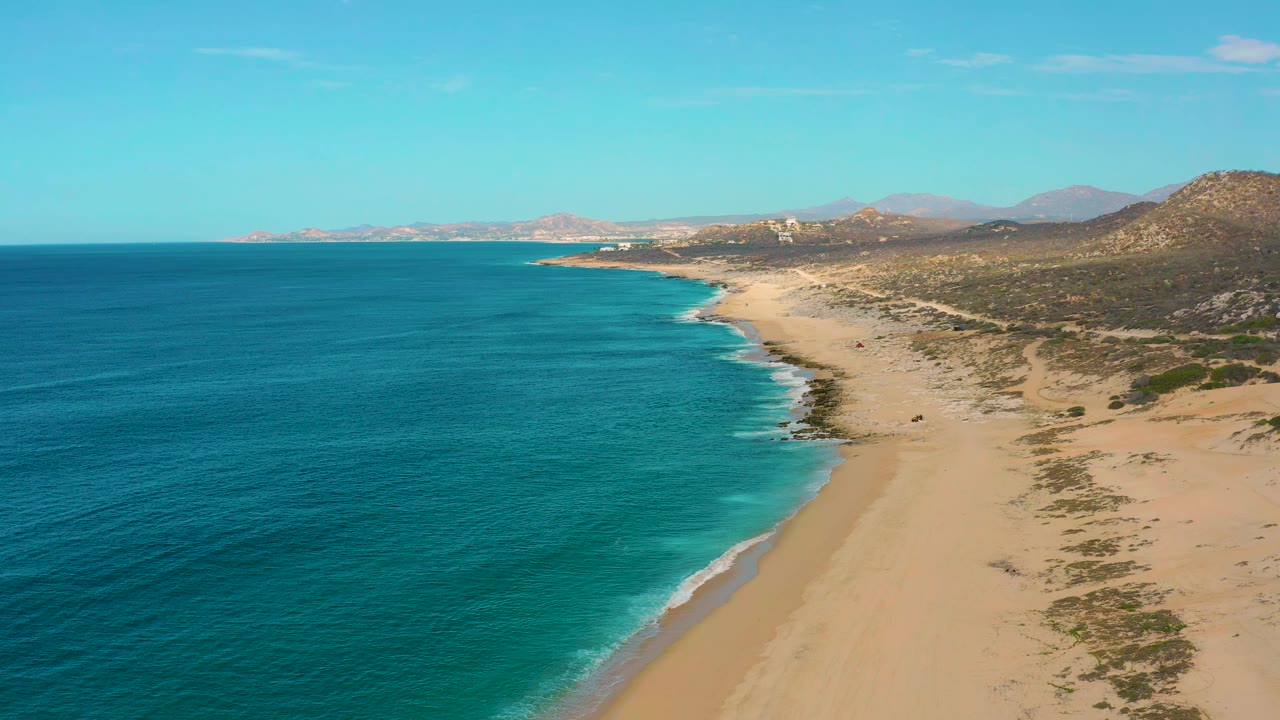 Laguna Beach coastline in Baja California Sur, Mexico, seen from above with the ocean filling the left side, a long empty sand strip at center, rocky hills at right, and no visible people.