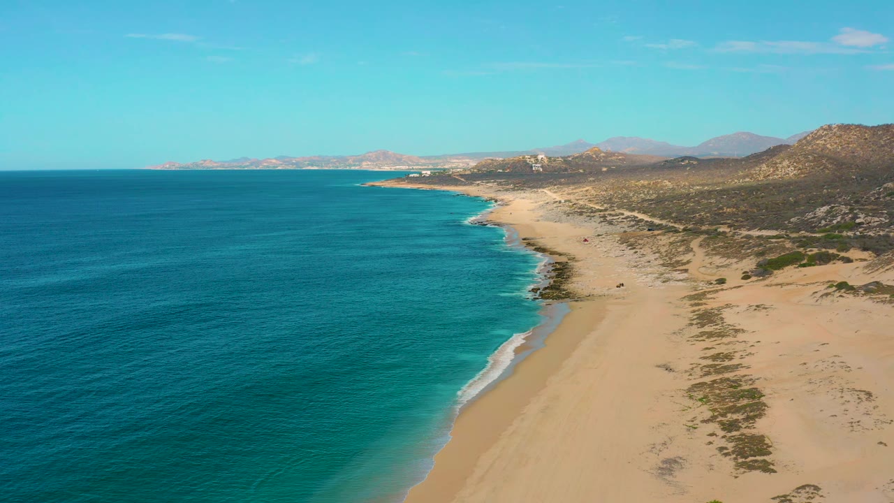 Laguna Beach aerial view showing a broad band of blue-green ocean, a narrow white surf edge, pale sand, inland scrub, distant hills, and no visible people.