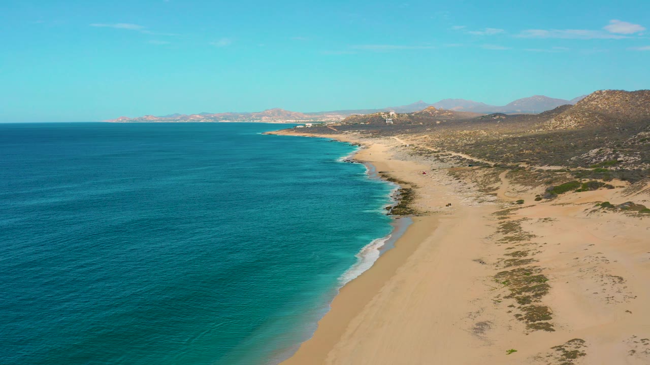 A drone-height frame of Laguna Beach with darker ocean water in the foreground, lighter turquoise shallows near shore, sandy coastline, hills beyond, and no visible people.