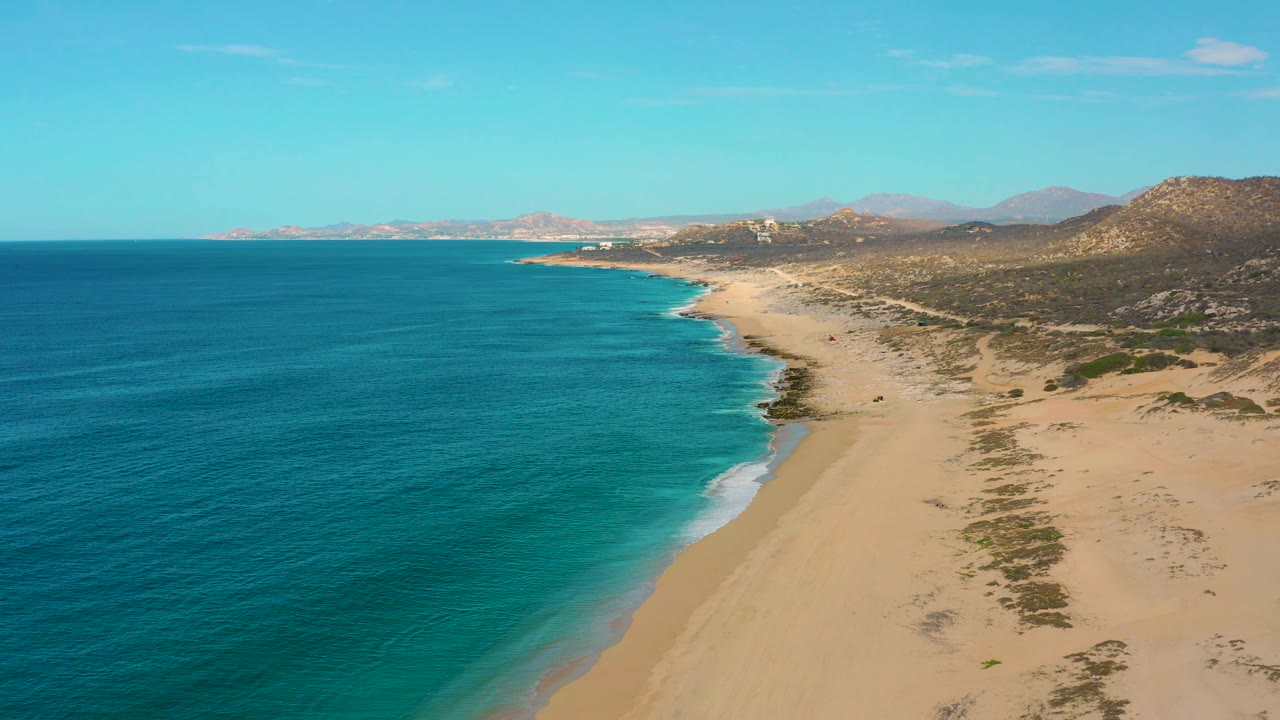 Wide aerial view over Laguna Beach with ocean occupying most of the left frame, a long empty sand strip curving into the distance, inland brush, and no visible people.