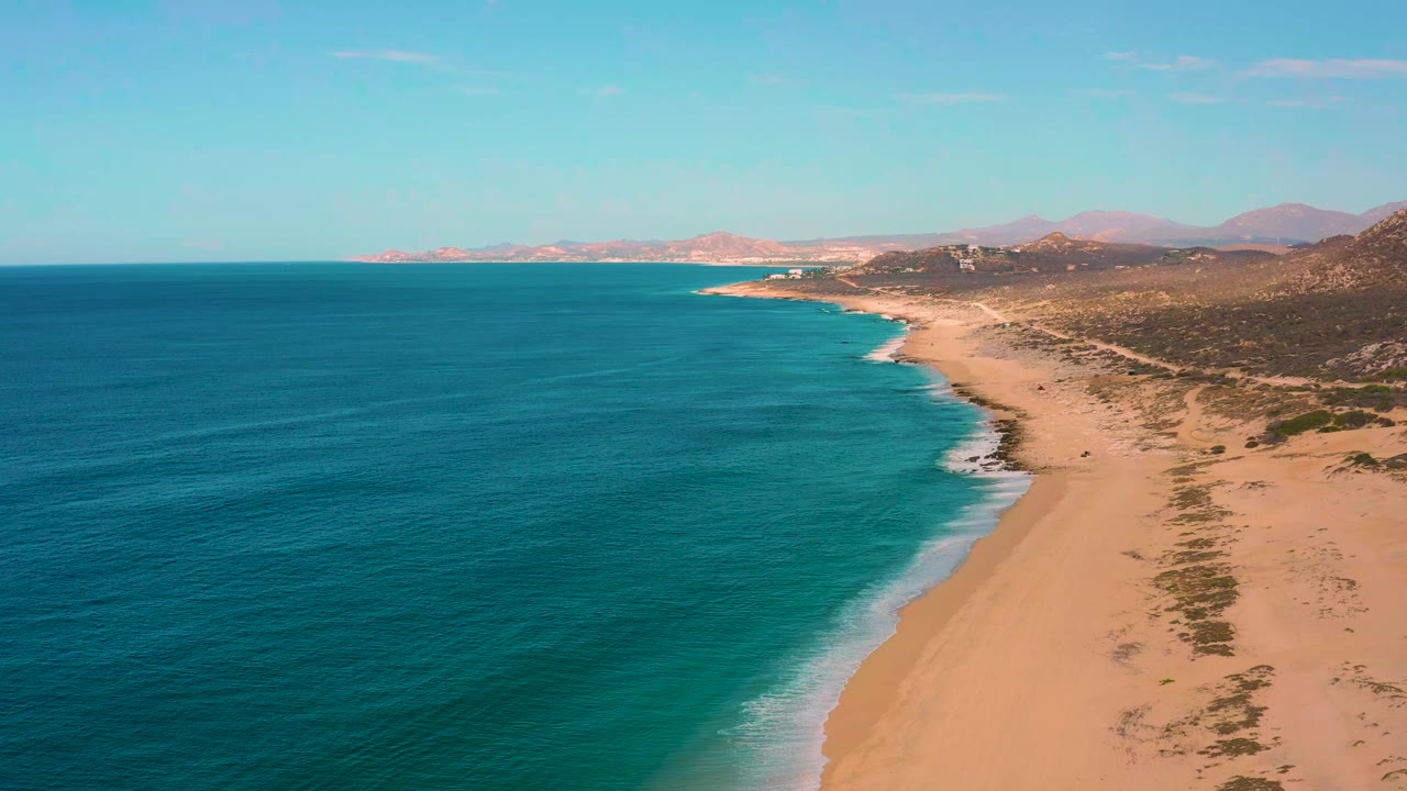 Laguna Beach shoreline in Baja California Sur with broad blue water, a thin breaking wave line, tan sand, sparse vegetation, far mountains, and no visible people.