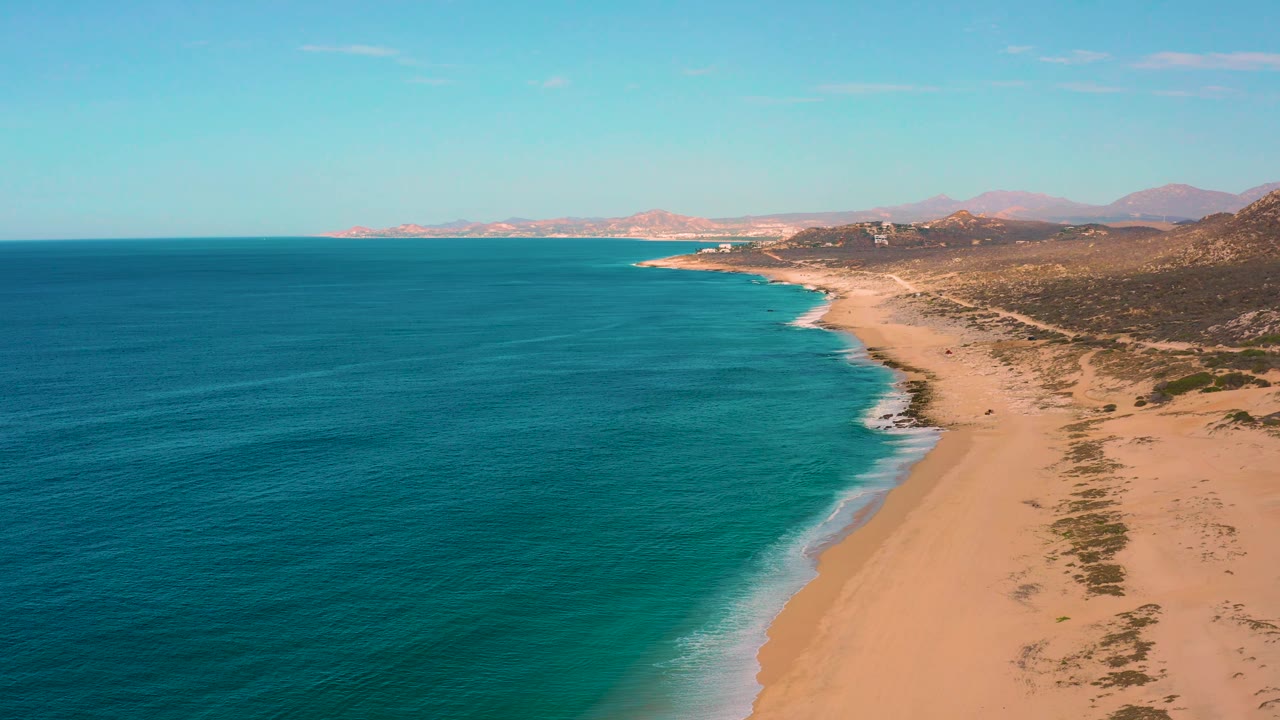 Aerial view over Laguna Beach, Baja California Sur, showing deeper blue water in the foreground, lighter water near shore, empty sand, low scrub, distant ridges, and no visible people.