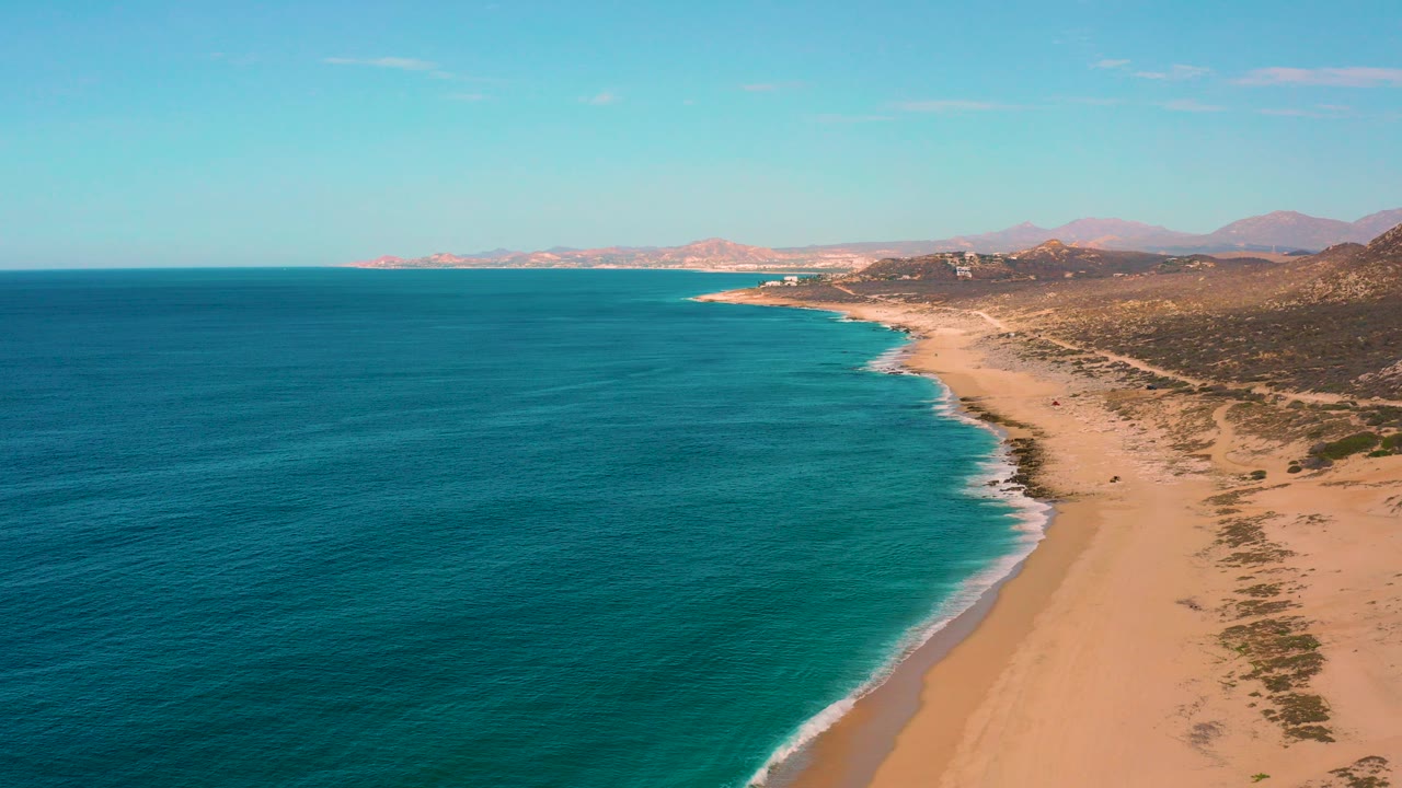 Aerial frame over Laguna Beach, Baja California Sur, showing blue ocean on the left, a long sandy shoreline on the right, low hills in the distance, and no visible people.