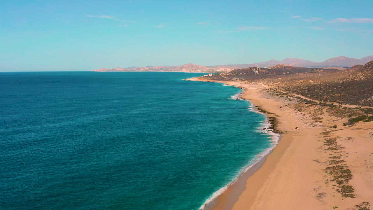 Aerial view of Laguna Beach, Baja California Sur, with turquoise water curving along the beach, scrub-covered land beyond the sand, distant mountains, and no visible people.