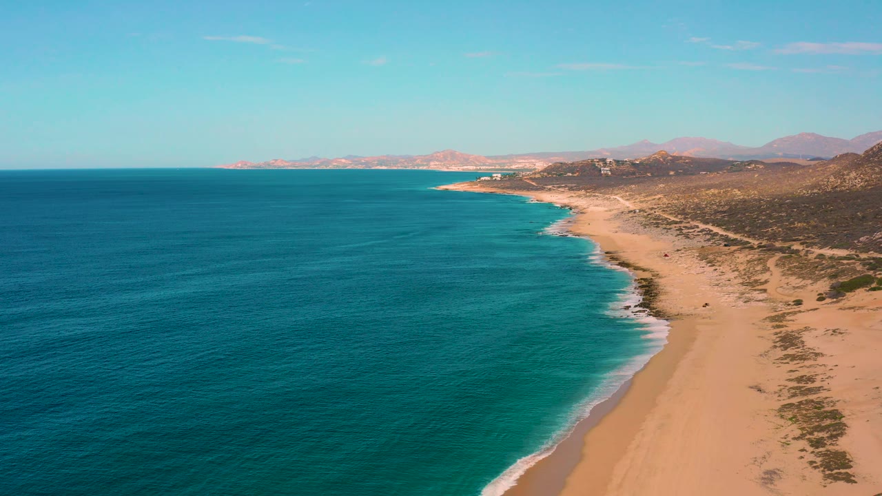 Aerial frame from Laguna Beach, Baja California Sur, showing textured blue water, a pale sand beach, dry coastal vegetation, a low mountain backdrop, and no visible people.