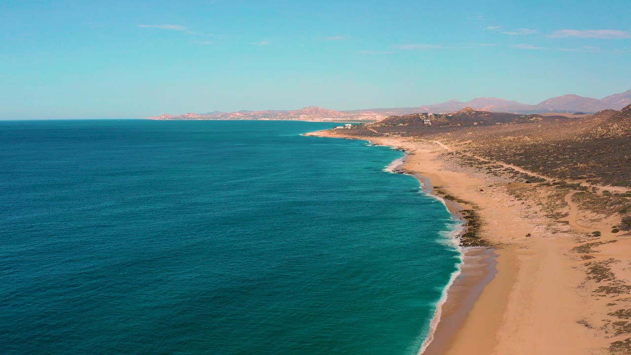 Opening aerial frame over Laguna Beach, Baja California Sur, with deep blue ocean on the left, a sandy shore on the right, distant low hills, and no visible people.