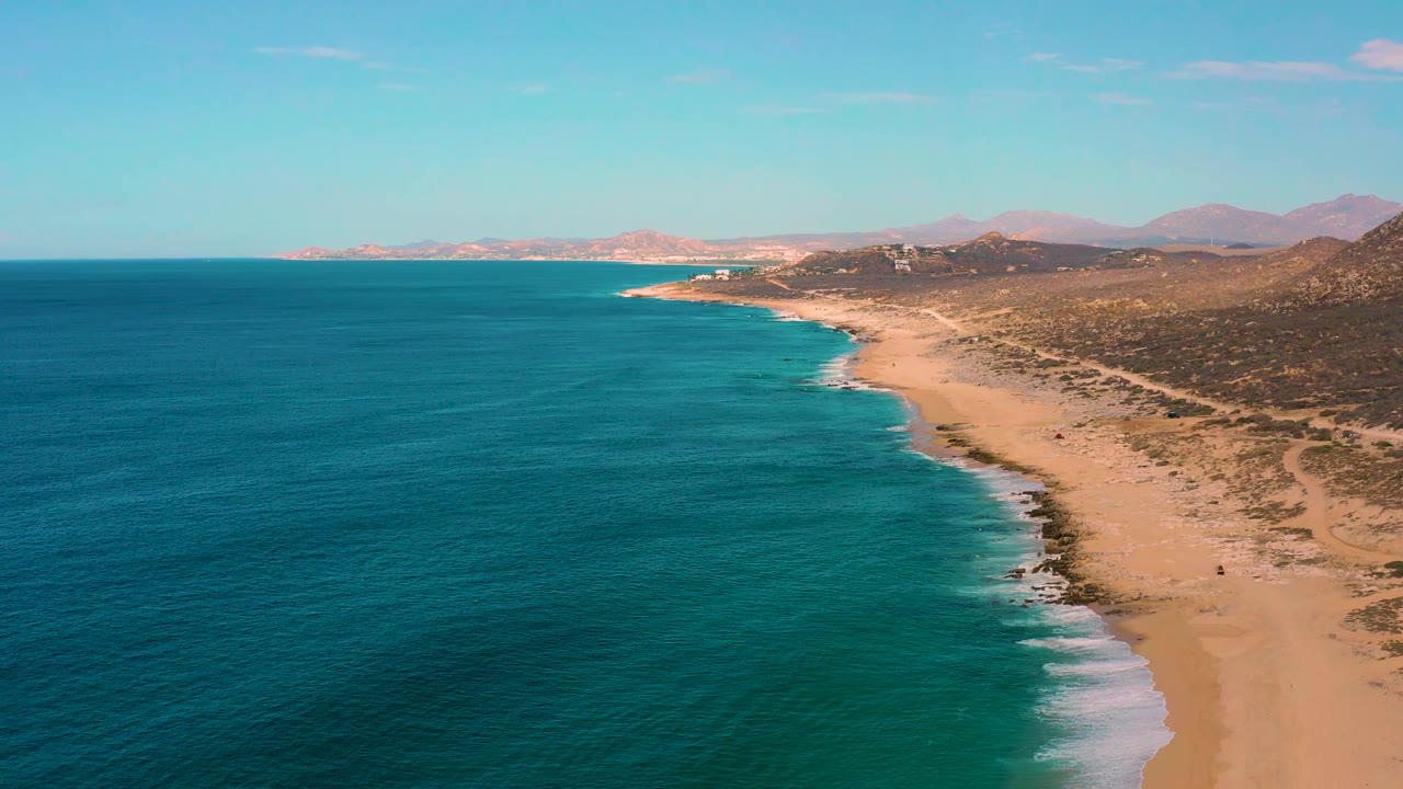 Aerial view along Laguna Beach in Baja California Sur with broad blue-green ocean, a thin line of white surf, dry coastal terrain, distant mountains, and no visible people.