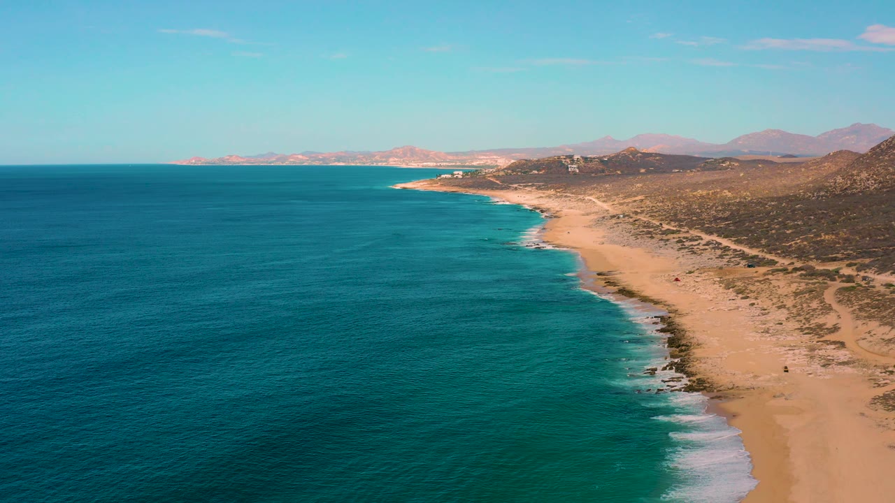 High aerial frame of Laguna Beach, Baja California Sur, with open teal water across most of the image, sandy beach at right, low scrubland beyond it, and no visible people.