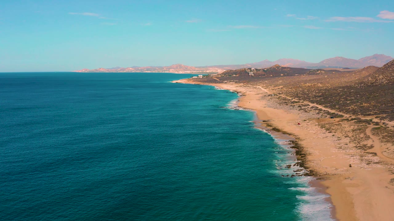 Aerial frame over Laguna Beach, Baja California Sur, showing the sandy coast running into the distance, calm blue water on the left, pale surf at the waterline, and no visible people.