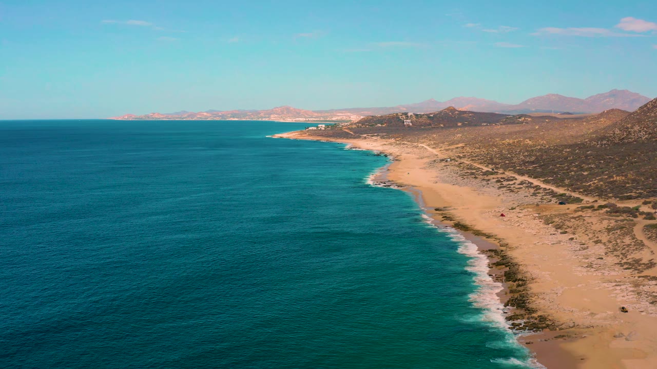 Laguna Beach coastline from above, showing the dark blue ocean widening at left, a pale sand beach edged by white waves, brown hills inland, and no visible people.