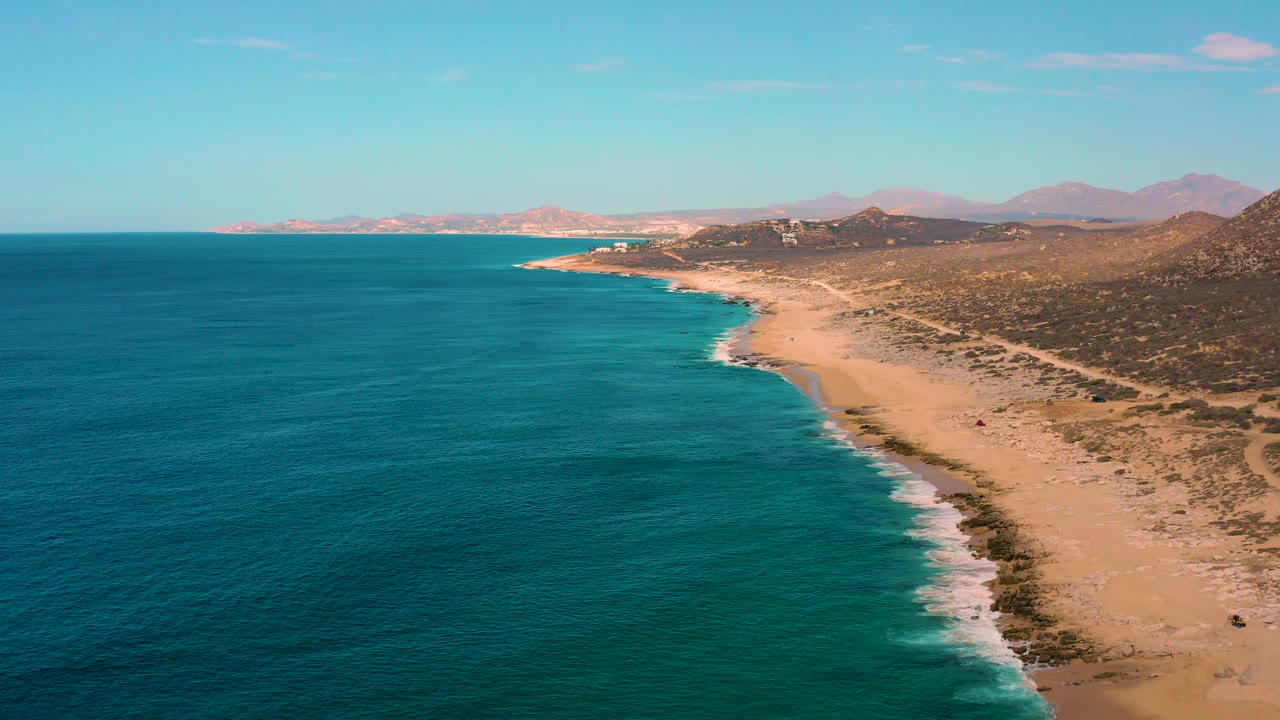 Aerial frame over Laguna Beach with deep blue water in the foreground, turquoise shallows near the surf line, a long sandy beach, inland hills, distant mountains, and no visible people.