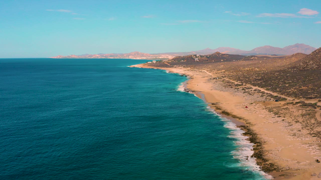 Laguna Beach aerial view with foamy waves tracing the sand, the ocean occupying most of the left side, scrub-covered hills on the right, distant mountains, and no visible people.
