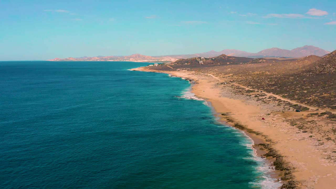 Aerial view of Laguna Beach in Baja California Sur, Mexico, showing blue-green water filling the left side, a thin white surf edge, tan beach, brushy terrain, distant mountains, and no visible people.