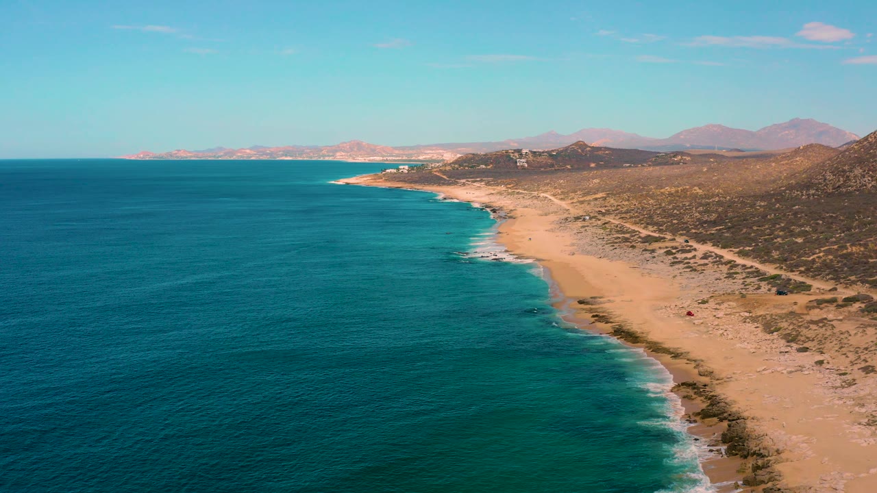 Laguna Beach aerial frame in Baja California Sur, Mexico, with the ocean taking most of the left foreground, the sandy beach bending toward the distance, dry brush and hills to the right, and no visible people.
