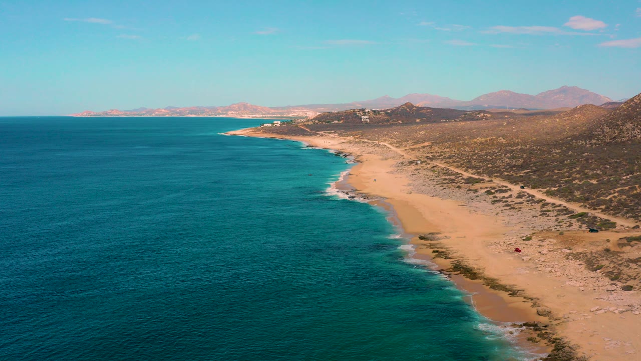 Aerial view over Laguna Beach, Baja California Sur, with blue sea filling the left side, a sandy shoreline curving right, distant mountains, and no visible people.
