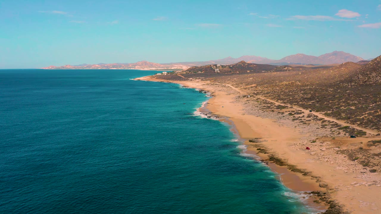 Laguna Beach coastline seen from above, with turquoise water near shore, deeper blue sea offshore, tan sand, scrub-covered land, distant mountains, and no visible people.