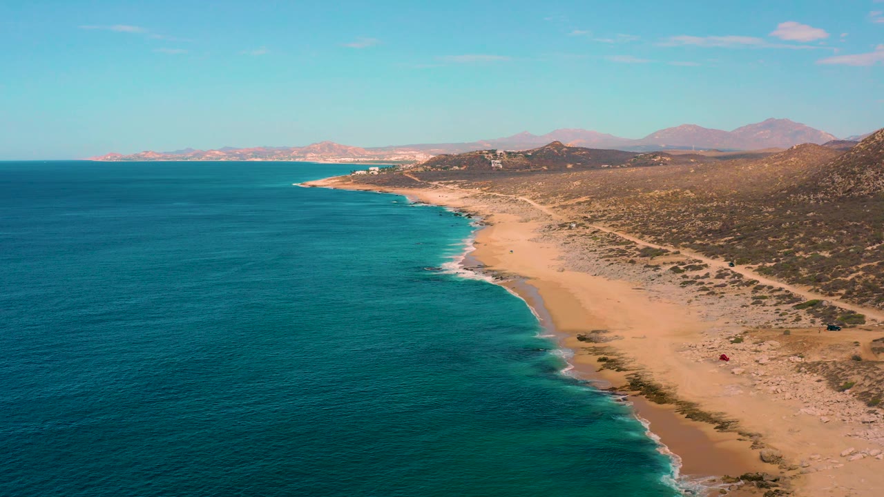 Wide overhead view of Laguna Beach, Baja California Sur, where broad blue water meets a pale sandy coast, with scrubland and mountain ridges beyond, and no visible people.