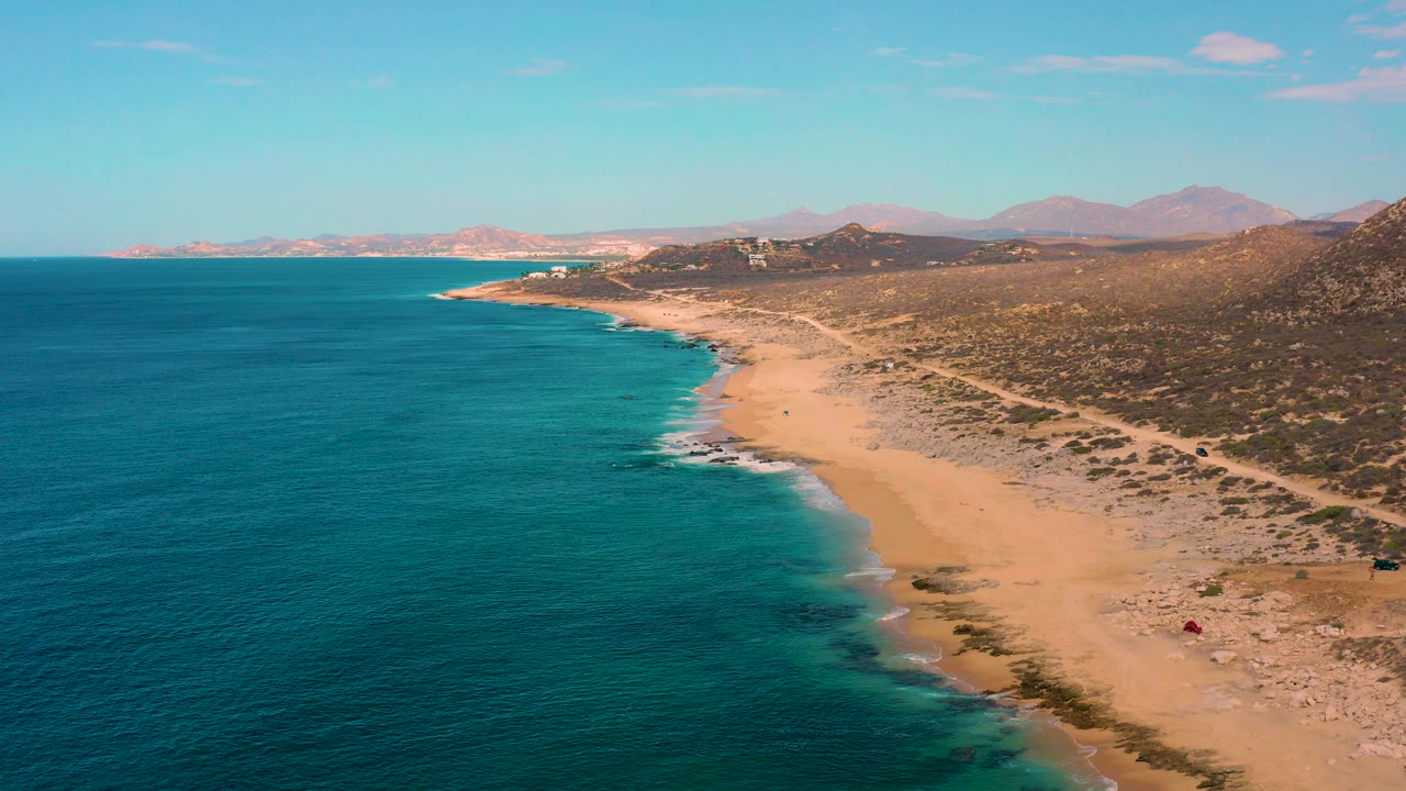 A wide aerial view over Laguna Beach, Baja California Sur shows turquoise ocean on the left, a long sandy beach on the right, dry hills beyond the shore, and no visible people.